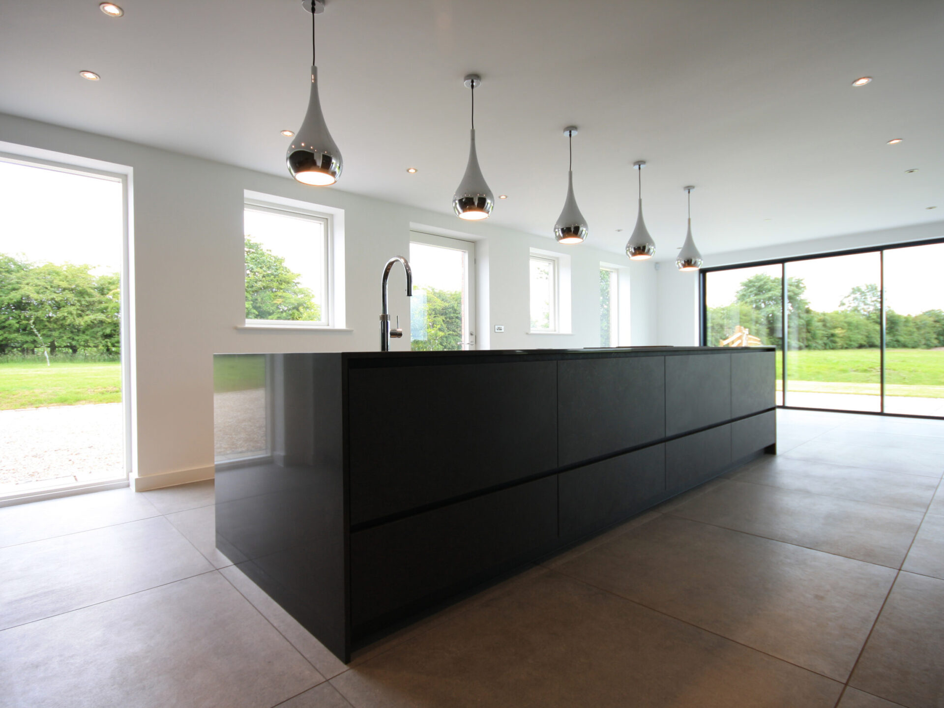 black kitchen units with chrome pendant lights with tiles flooring, windows and doors looking out onto surrounding green spaces