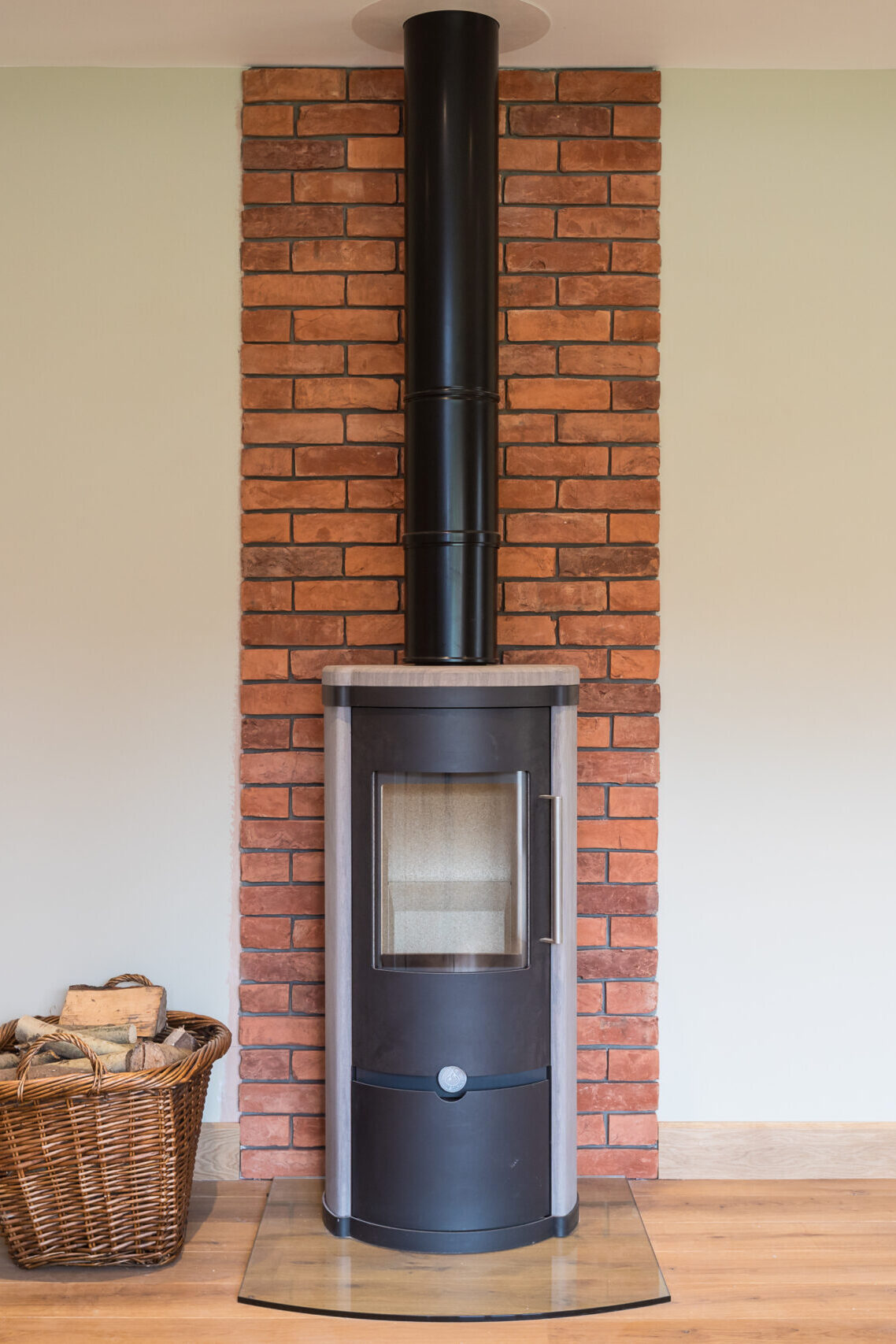 cylindrical wood burning stove with fire wood basket and brick wall feature behind
