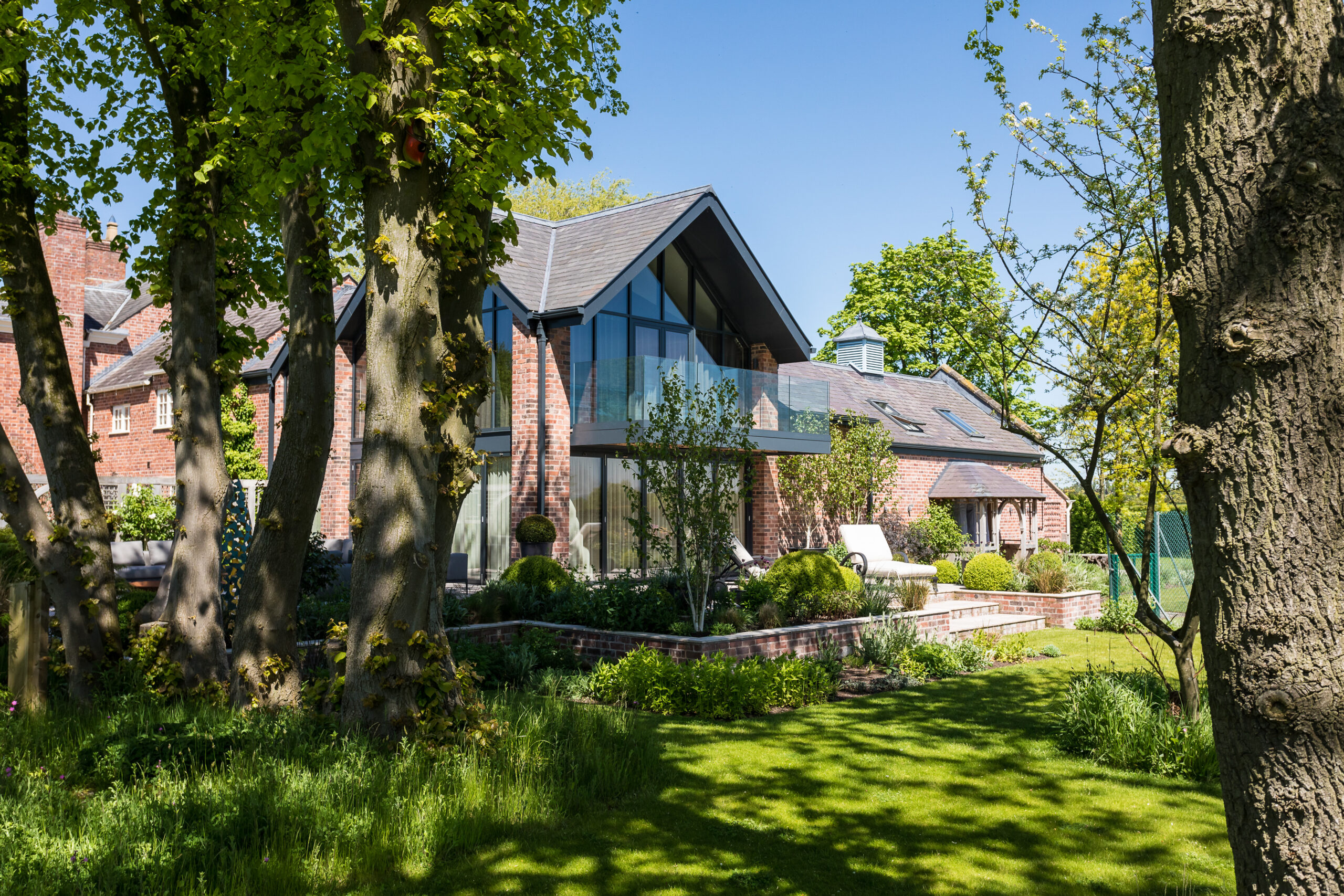 Ash House exterior view through trees of Ash House extensions with external balcony and gable end glazing