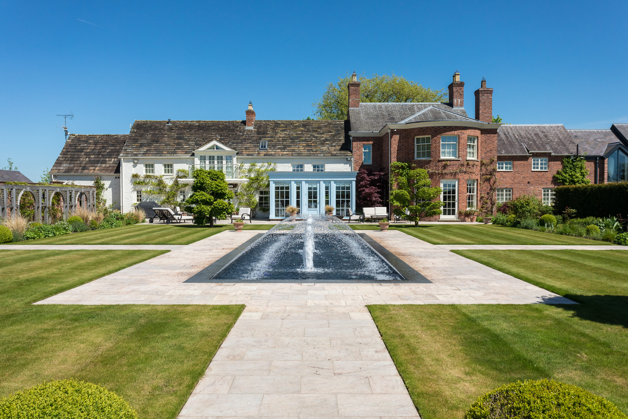 view of Ash House from across water fountain feature and courtyard with paved path and curated planting with blue garden room extension