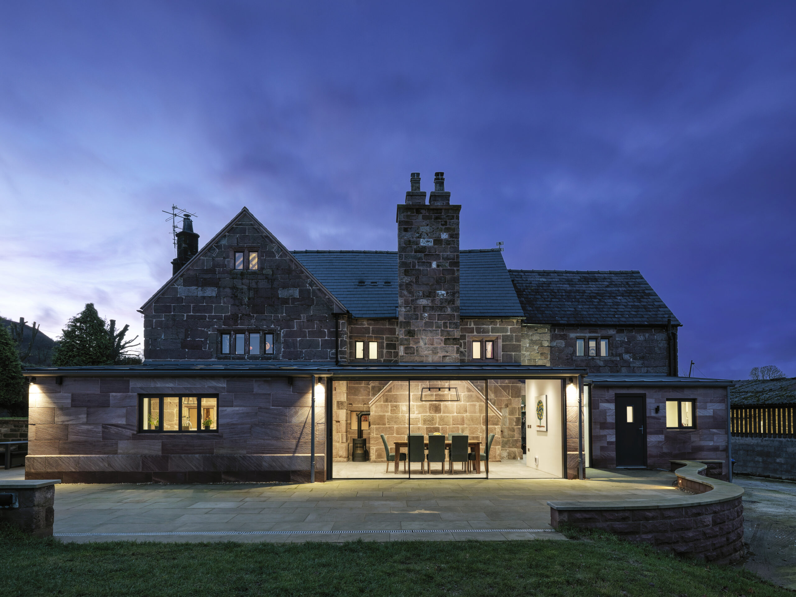 Evening view of a new single storey rear extension to a Grade 2 listed farmhouse with large triple pane full height sliding doors framing views into the space with the renovated stone chimney being highlighted by artificial lighting strategically placed.