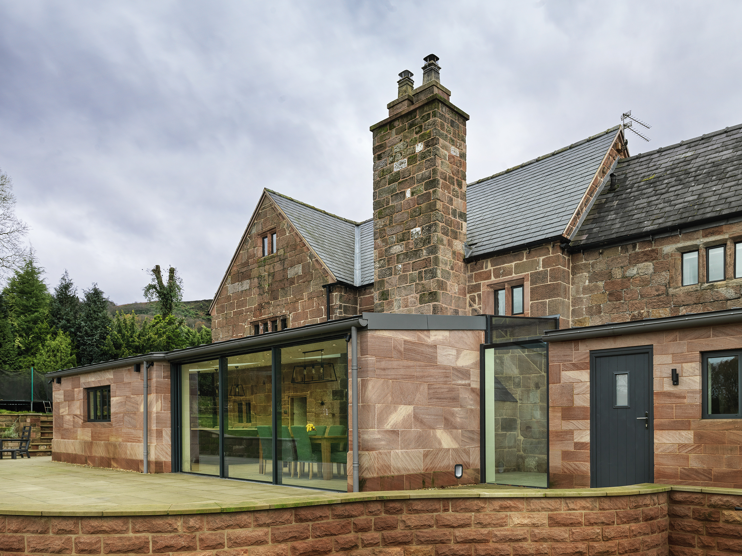 external view of a new rear extension to a Grade 2 listed farmhouse with coursed stone to complement the original stonework and stone mullion windows. Thin framed openings, roof glazing and zinc roof covering creates a modern, sleek extension