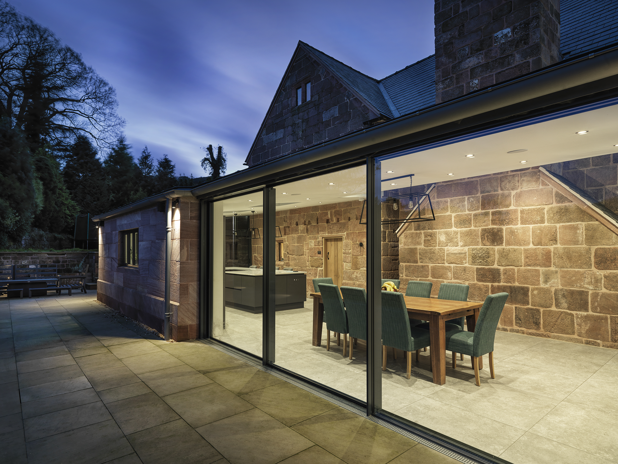 evening shot of a rear extension to a Grade 2 listed farmhouse with large triple pane floor to ceiling sliding doors in the dining area with spotlights highlighting the original stonework walls and chimney.